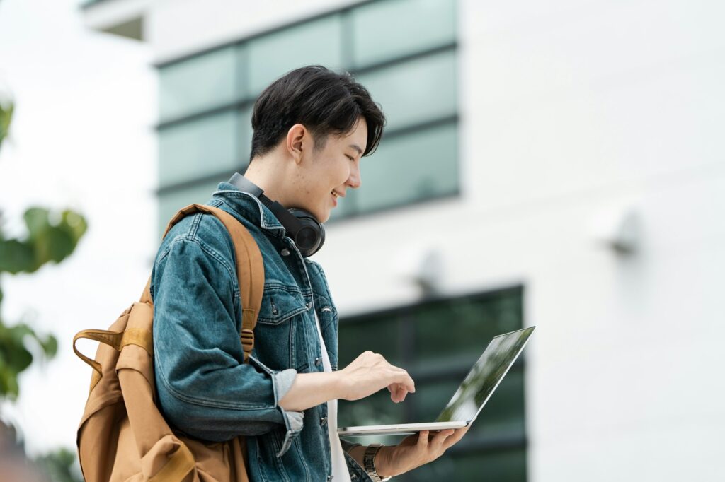 Young Man Using Laptop Outdoors with Modern Building in Background, Wearing Denim Jacket and